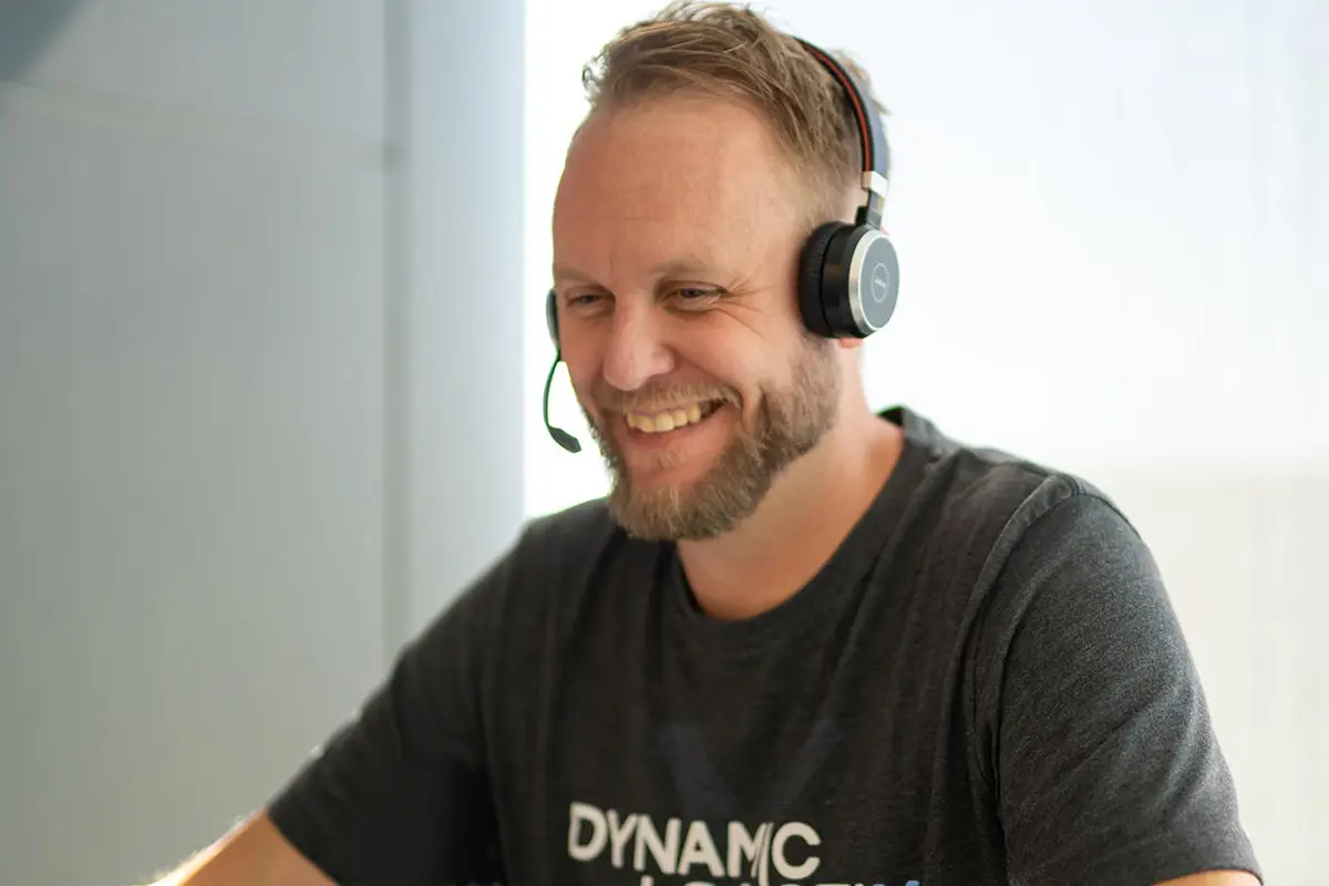 A smiling man with a beard wears a headset and a dark DYNAMIC shirt while sitting at a desk, appearing to be engaged in a freight brokerage video or phone call.