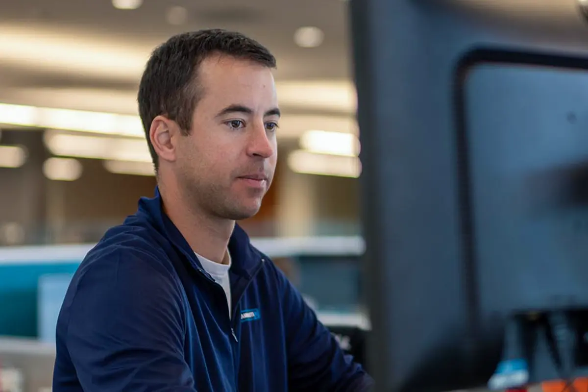 Man with short dark hair wearing a blue jacket sits at a desk, looking intently at a computer monitor in a modern office setting, focused on managed transportation solutions.
