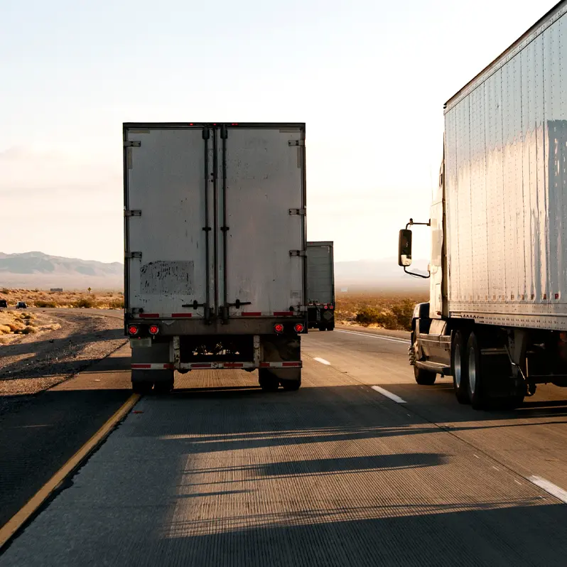 Two semi-trucks travel down a multi-lane desert highway at sunset, leaving long shadows on the road ahead.