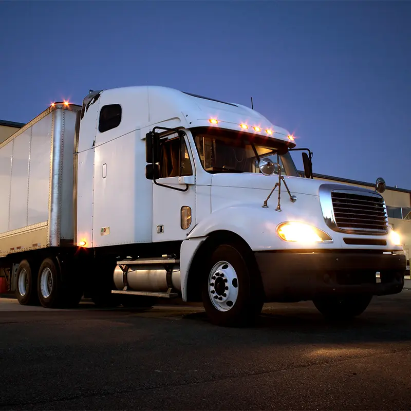 A white semi-truck with its lights on, belonging to meat shipping experts, is parked on a paved surface at dusk, with a building visible in the background.