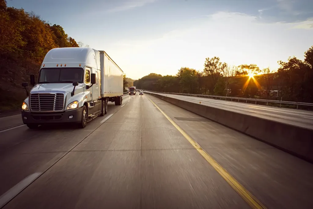 A white semi-truck, cleared through carrier vetting, drives on a highway during sunset, surrounded by trees and other vehicles. The sun casts a warm light over the road and landscape.