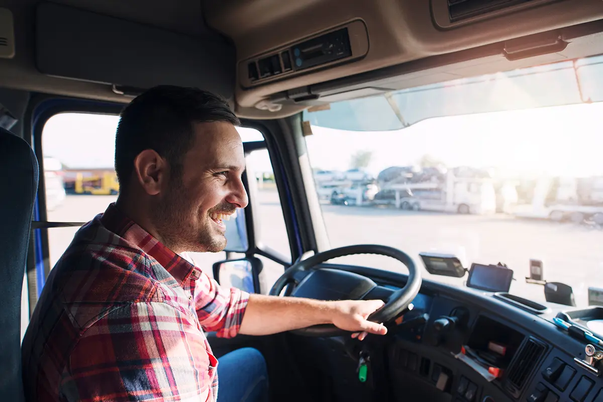 A smiling man in a plaid shirt drives a truck, holding the steering wheel with one hand. Sunlight streams through the window as vehicles and freight carrier resources fill the parking area outside.