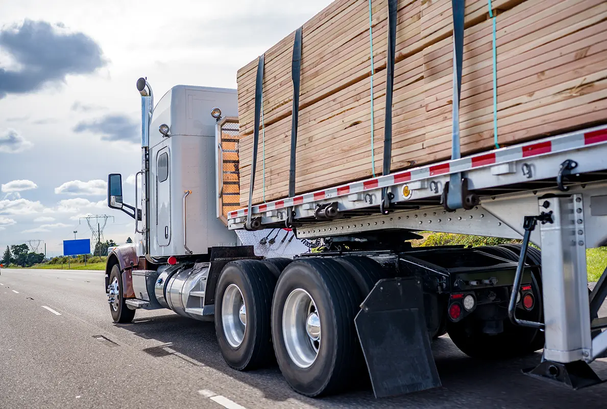 A semi-truck hauls a flatbed trailer loaded with neatly stacked lumber, showcasing logistics solutions by industry as it travels on a highway under a partly cloudy sky.