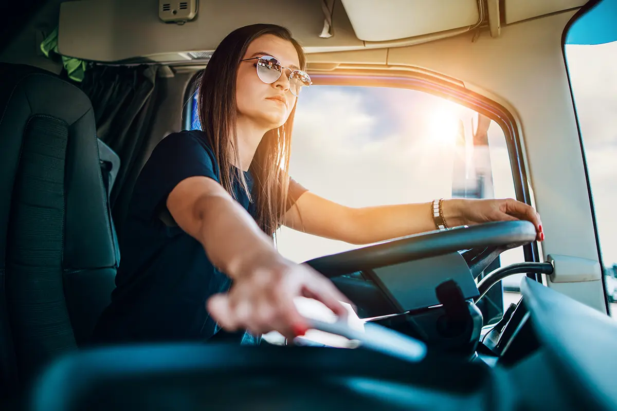 A woman wearing sunglasses drives a truck, holding the steering wheel with one hand and reaching for a control with the other, sunlight streaming through the window—confidently managing her freight carrier resources.