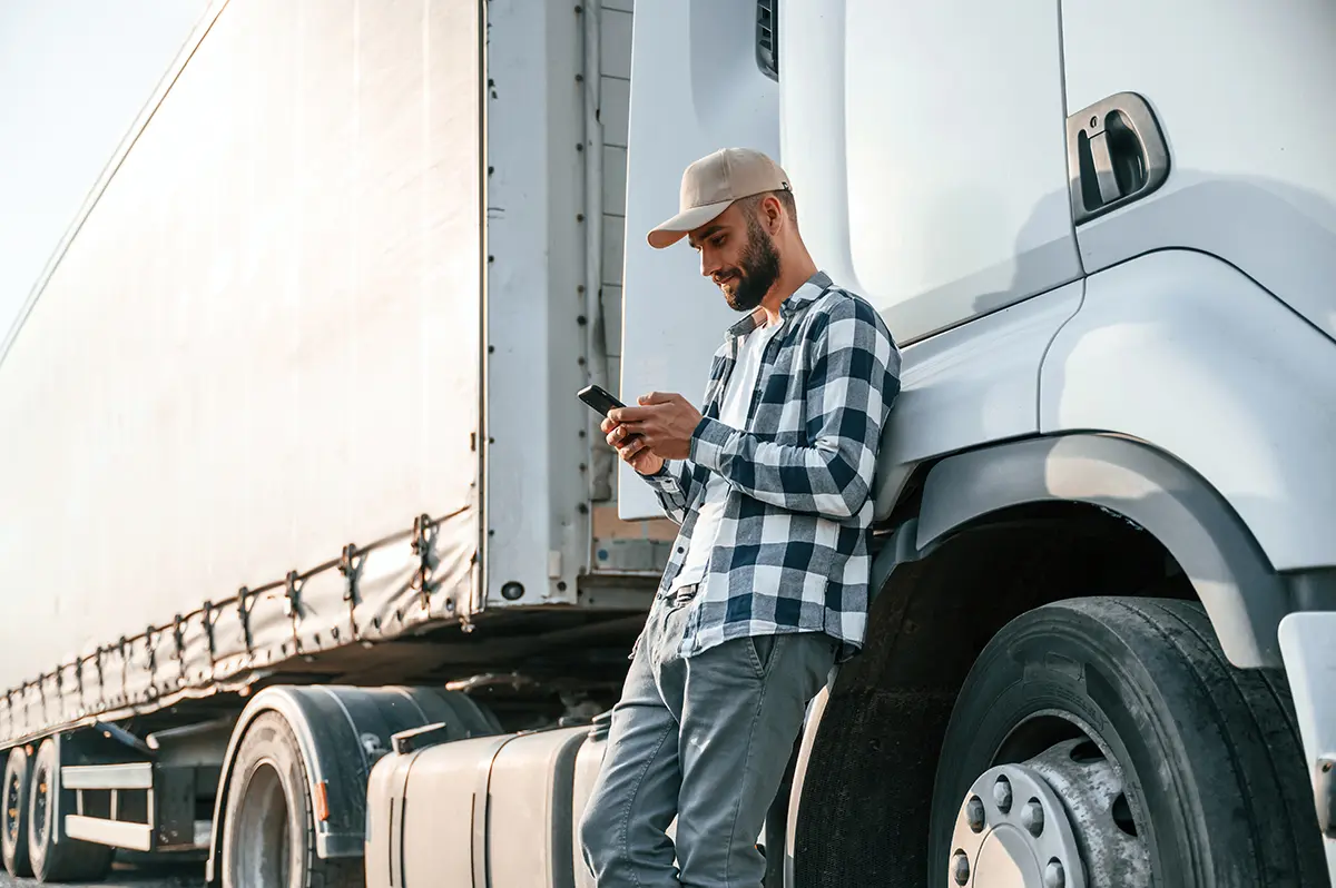 A man in a plaid shirt and cap leans against a white semi-truck, smiling while looking at his smartphone as he checks freight carrier resources.