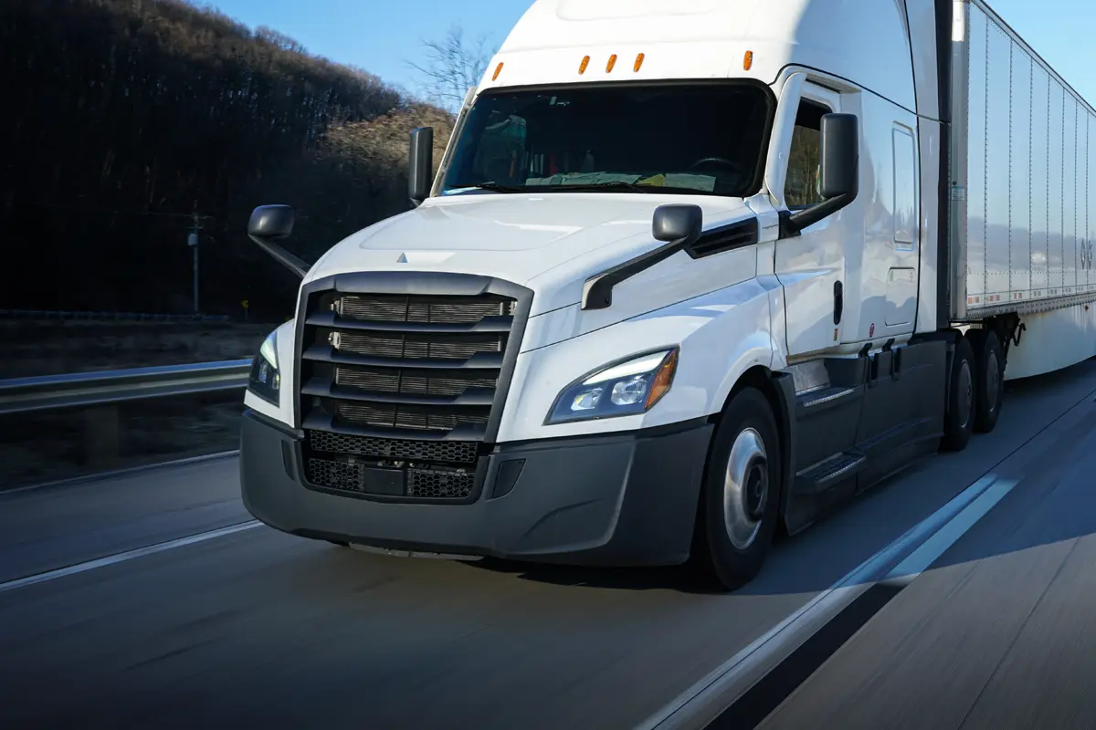 A white semi-truck with a trailer, part of top freight carrier resources, drives down a highway in daylight, passing trees and barriers, with the road and sky visible in the background.