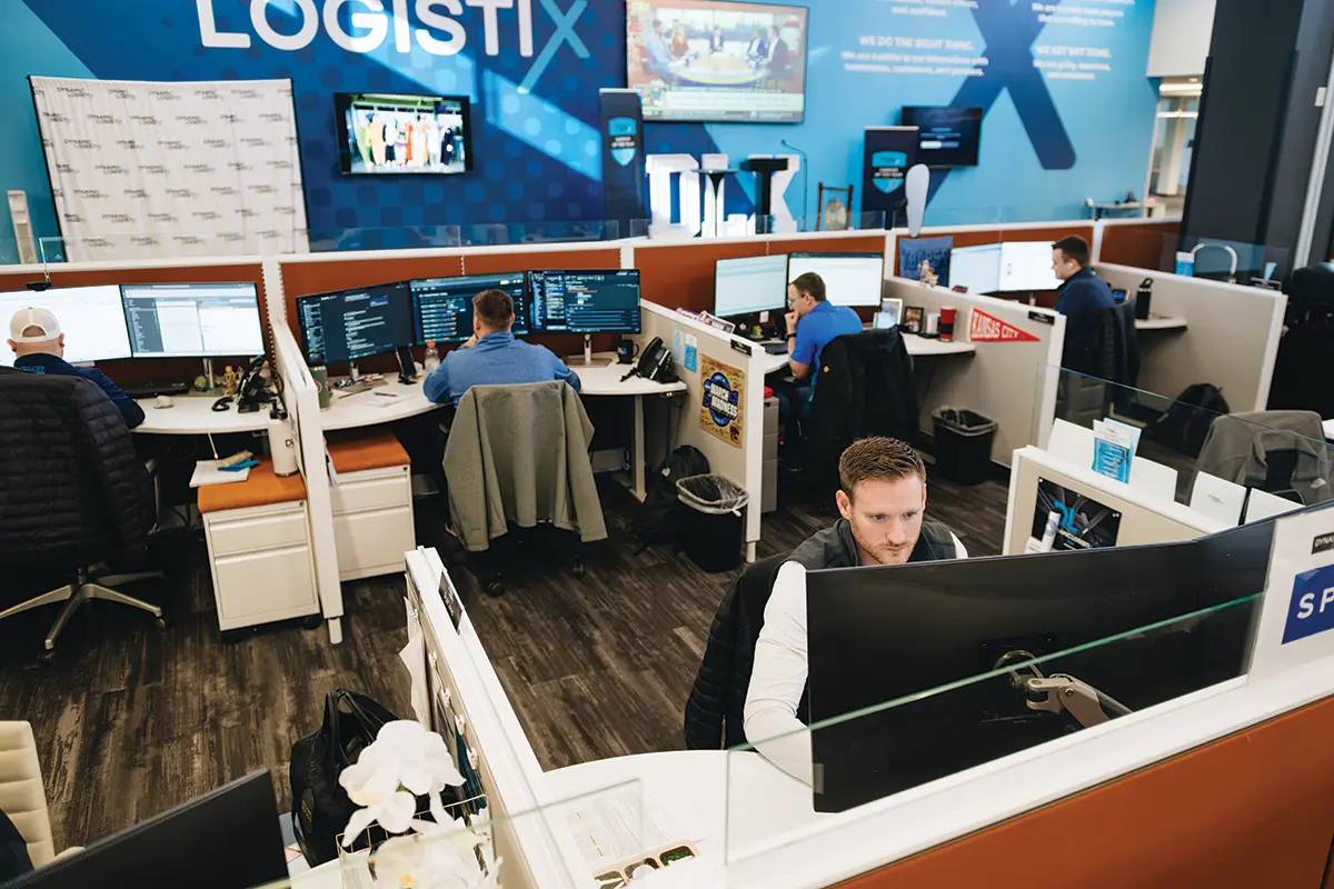 Office space with several people working at computer desks. Multiple monitors, modern decor, and blue LOGISTIX branding highlight the busy, collaborative environment focused on logistics support services.