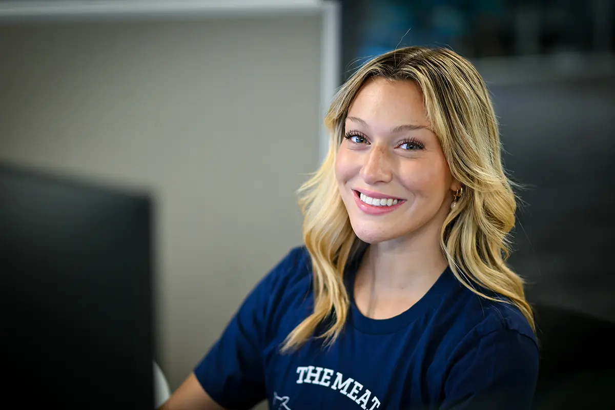 A smiling woman with wavy blonde hair sits at a desk, wearing a navy blue T-shirt. She looks toward the camera in a brightly lit, modern office specializing in freight bill audit services.