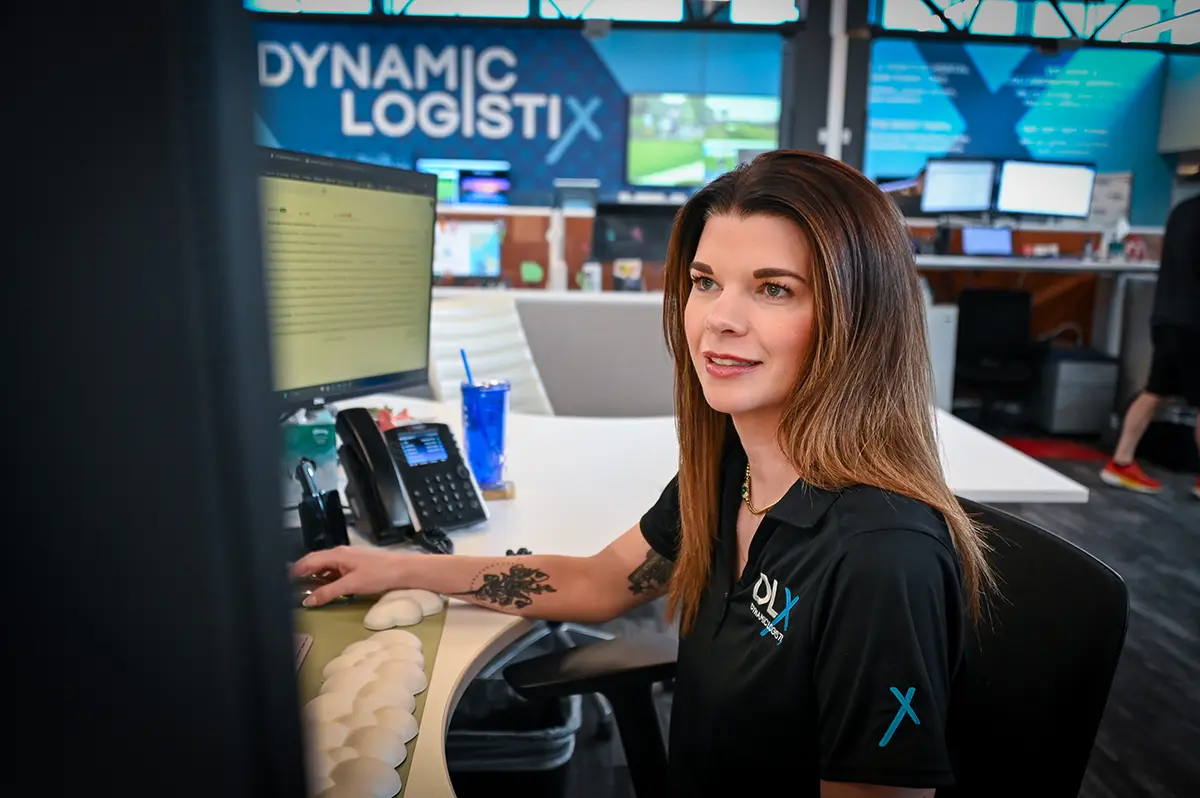 Woman with long brown hair sits at an office desk, working on a computer. She wears a black polo shirt and smiles, surrounded by office equipment and logistics KPIs. Dynamic Logistix is visible in the background.