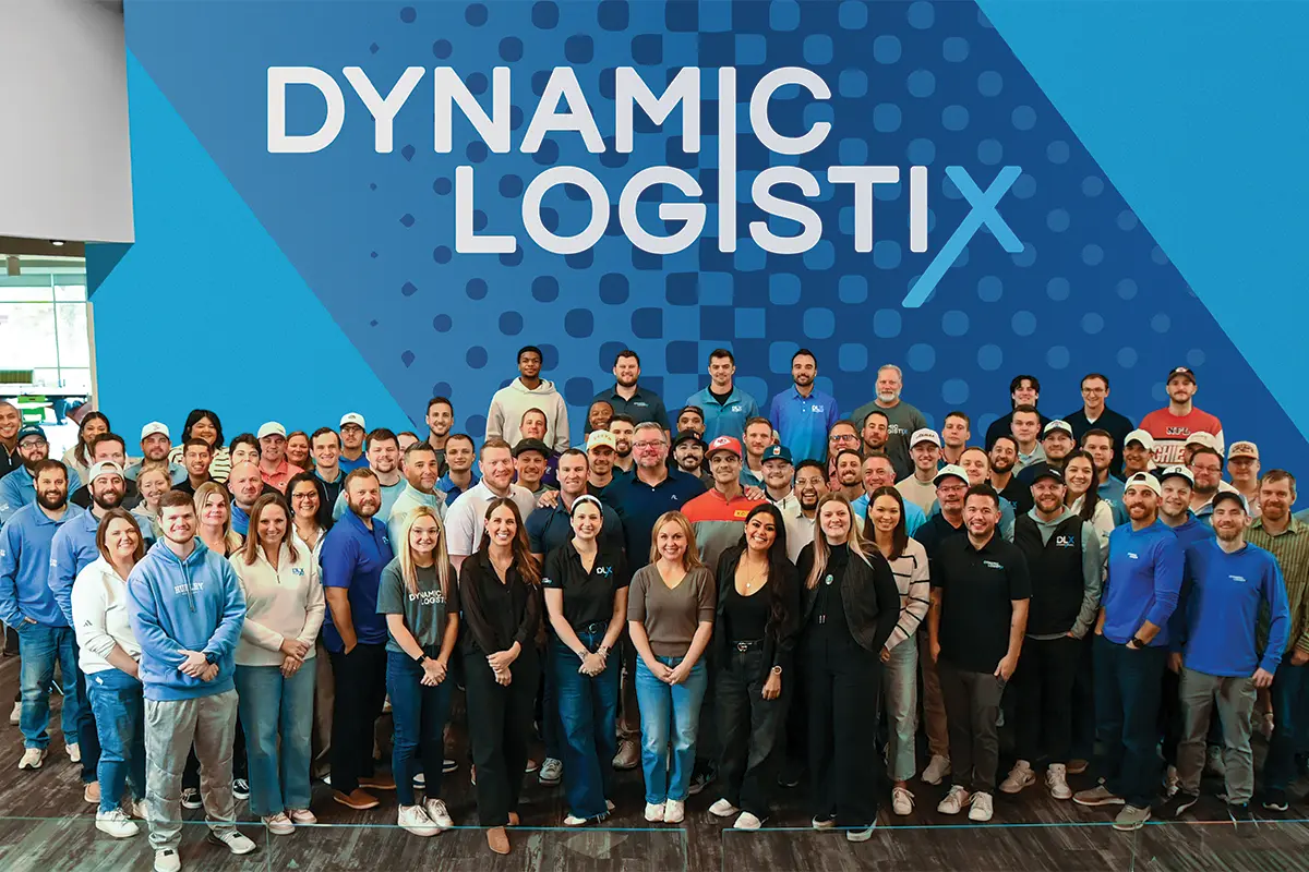 A large group of people poses and smiles together in front of a blue wall with the words Dynamic Logistix, a managed transportation company, written on it.