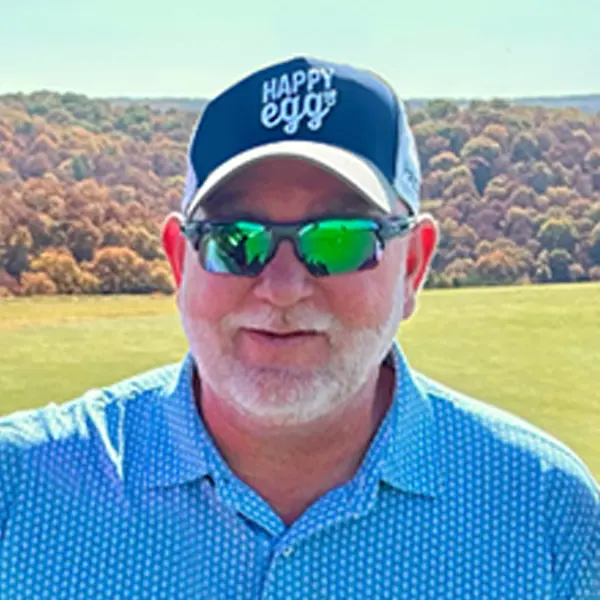A man with a white beard wearing sunglasses, a blue Happy Egg DLX cap, and a blue patterned shirt stands outdoors with green fields and trees in the background.