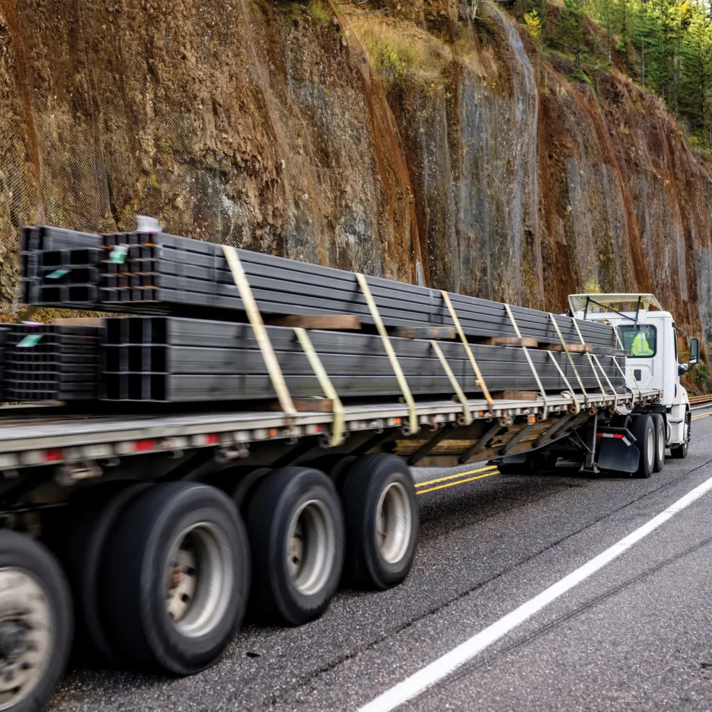 A white semi-truck transports a flatbed trailer loaded with bundles of long, dark metal beams secured by straps. The truck travels along a two-lane highway next to a steep, rocky hillside covered in protective mesh.