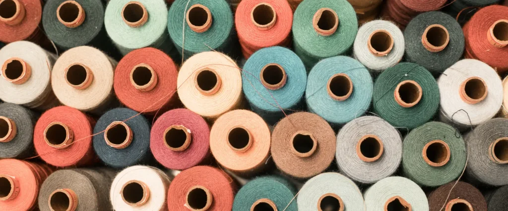 A high-angle, close-up view shows many spools of thread in various colors, including shades of blue, green, red, and brown. Each spool has a hollow brown cardboard core at the center.