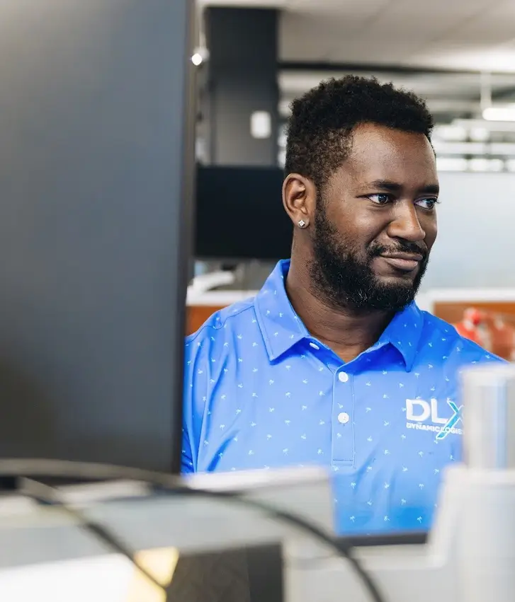A person with dark skin tone and a beard wears a blue polo shirt with a DLX Dynamic Logix logo while looking at a computer screen.