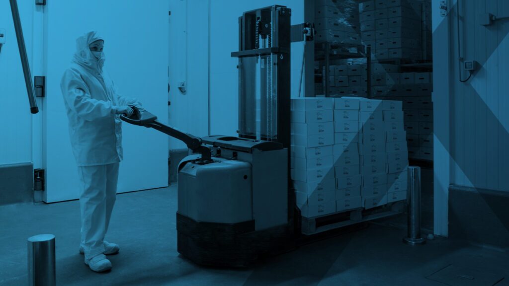 A person in protective clothing operates a pallet jack to move stacked boxes inside a storage facility with shelves and closed doors in the background.