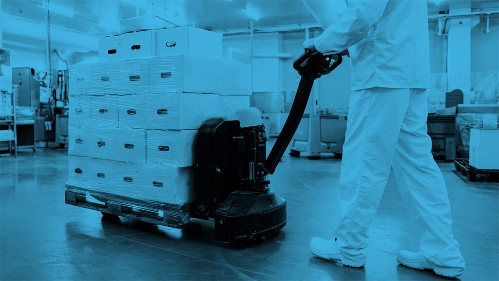 A person in white work clothes uses a pallet jack to move stacked boxes in a clean, industrial facility. The image is tinted blue.