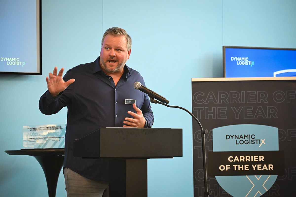 A man speaks at a podium with a microphone, gesturing with his hand. Behind him are signs and screens reading DYNAMIC LOGISTIX and CARRIER OF THE YEAR. A trophy is on a nearby table.