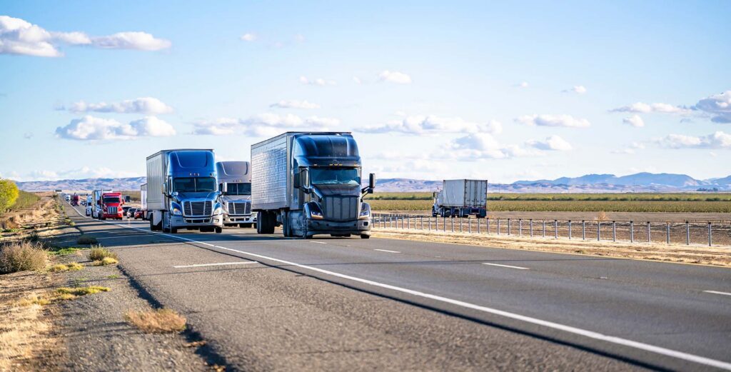 Several semi-trucks and trailers drive on a multi-lane highway through a dry, flat landscape with distant hills under a blue sky with scattered clouds.