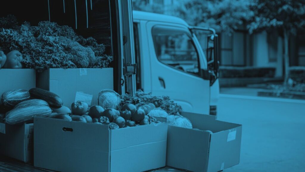 Boxes of assorted fresh vegetables are stacked on the back of a delivery van parked on a street, with another vehicle and trees visible in the background.