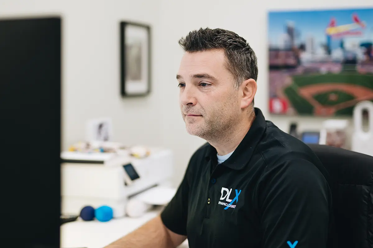 A person wearing a black polo shirt with a DLX logo looks intently at a computer monitor in an office setting.