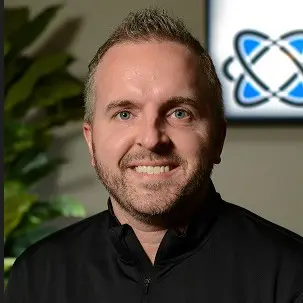 Smiling man with short hair and beard, wearing a black shirt, sits indoors with a plant and a blurred shipping technology logo featuring blue and black lines in the background.