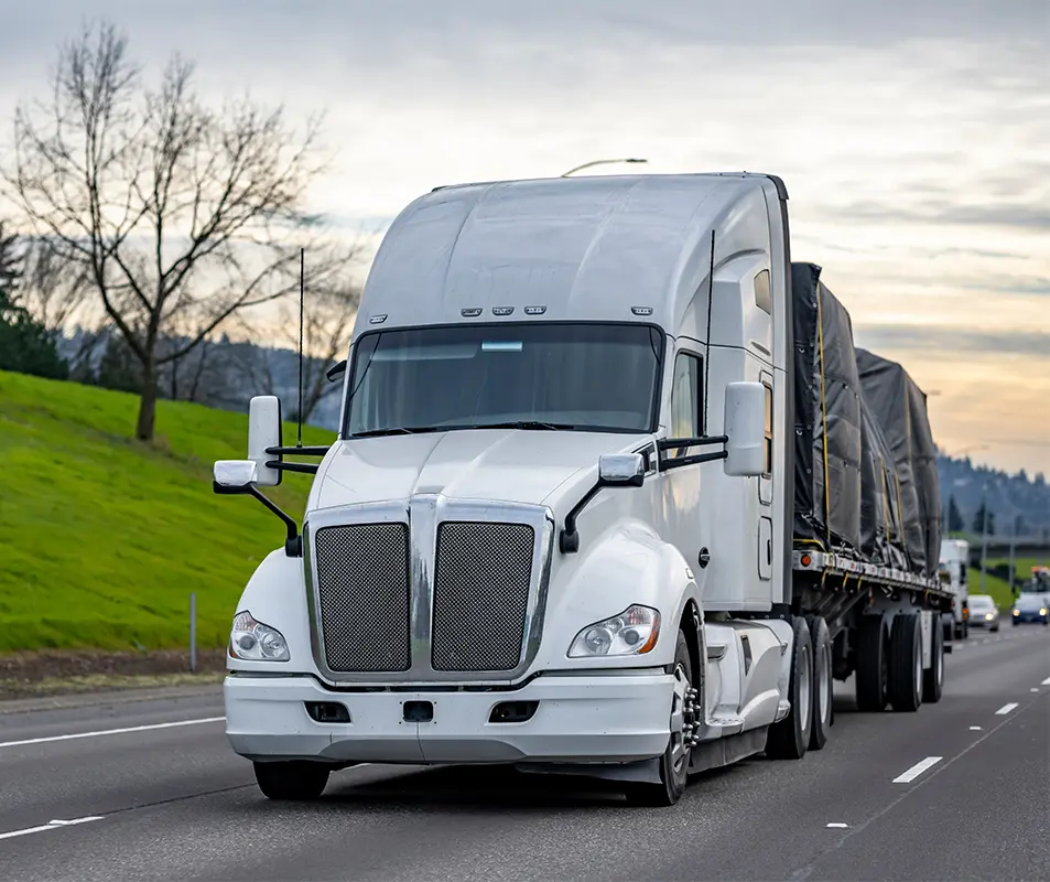 A white semi-truck with a covered flatbed trailer drives on a highway. The sky is cloudy, and there are grassy hills and bare trees visible along the roadside.