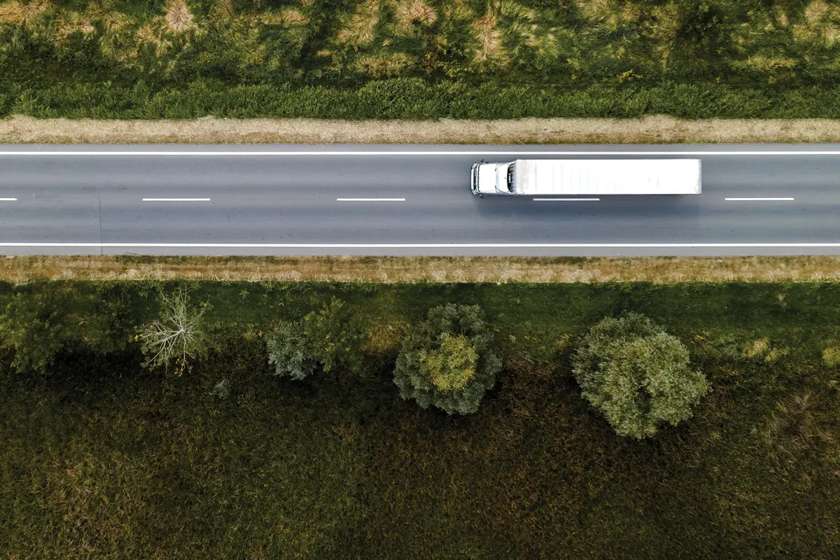Aerial view of a white semi-truck, powered by logistics technology, driving on a straight, empty road bordered by grass and trees on both sides.