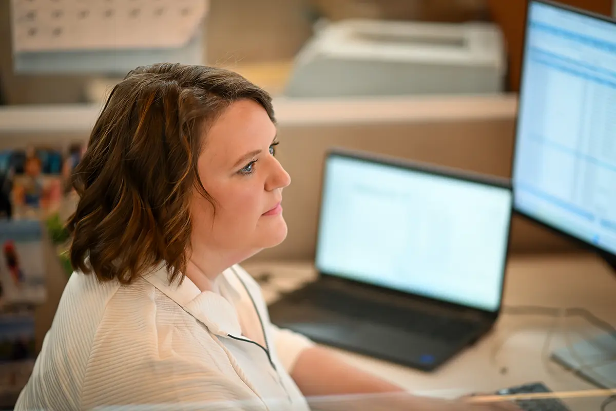 A woman with brown hair sits at a desk, focused on two computer monitors displaying shipping technology. She wears a white shirt; a laptop, papers, and office equipment are arranged around her.