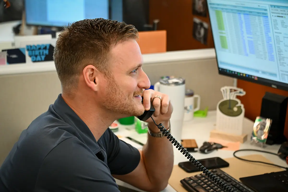 A man sits at his desk, smiling while talking on a corded phone. His desk holds a computer monitor displaying a logistics dashboard with spreadsheets, various office supplies, and a keyboard.