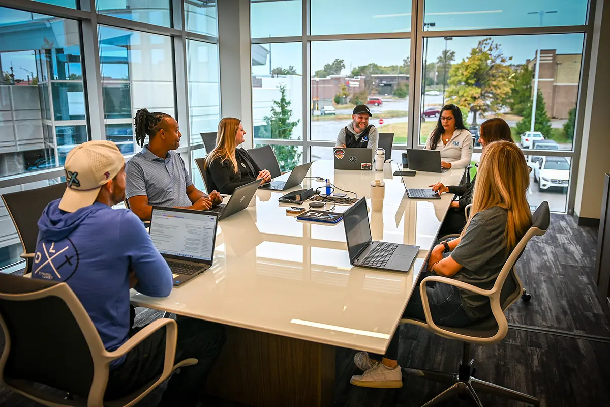 Eight people from the logistics support team sit around a conference table with laptops, engaged in discussion, in a bright office with large windows showing buildings and trees outside.