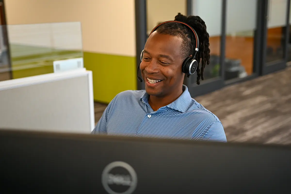 A man wearing a blue polo shirt and headphones smiles while working at a computer in a modern office specializing in specialized shipping.
