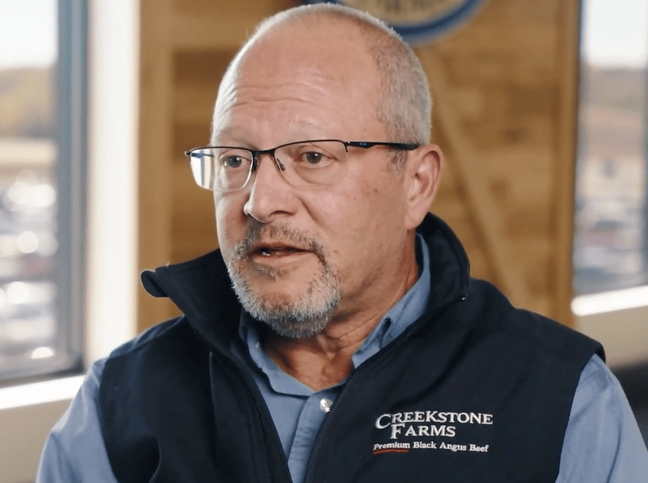 A man wearing glasses and a Creekstone Farms DLX Premium Black Angus Beef vest speaks indoors, with sunlight coming through windows in the background.