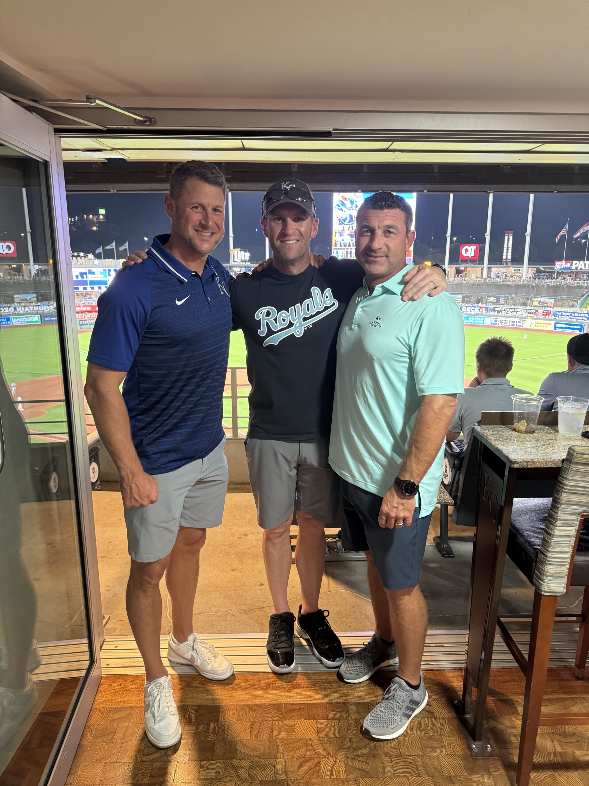Three men pose and smile together inside a stadium suite at a nighttime Kansas City Royals game, with polysource DLX signage and the bright field lights visible in the background.