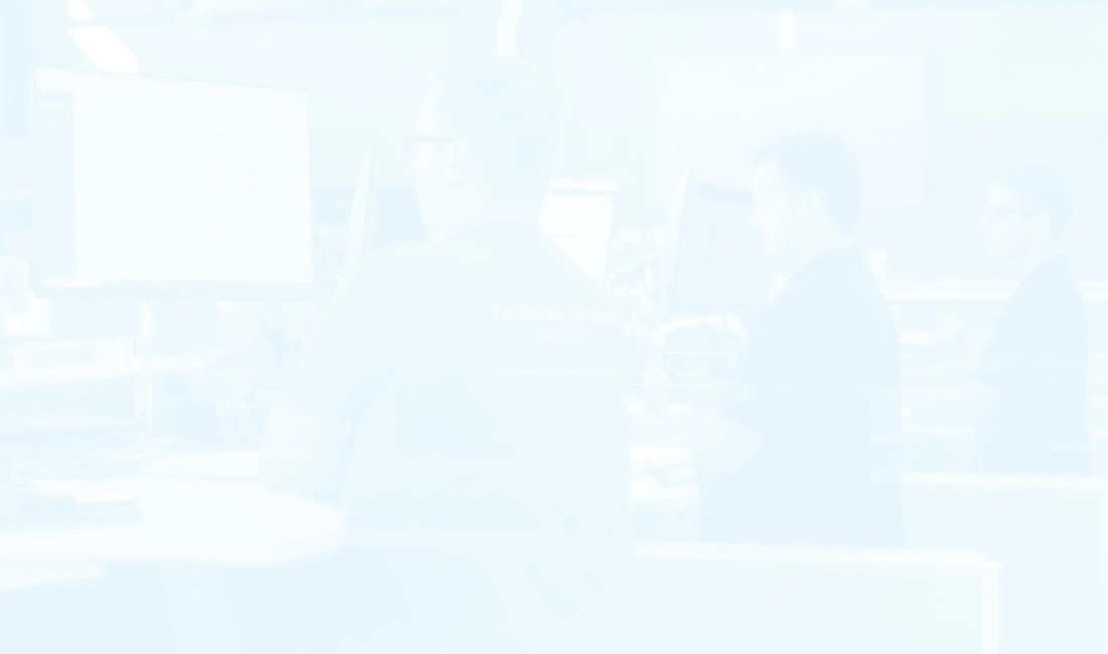 A faded, high-key photo of people working at computer desks in a bright, modern office setting.