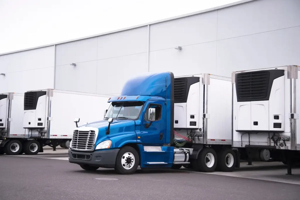 A blue semi-truck is parked in front of a large white warehouse, hitched to a white refrigerated trailer with its cooling unit visible.