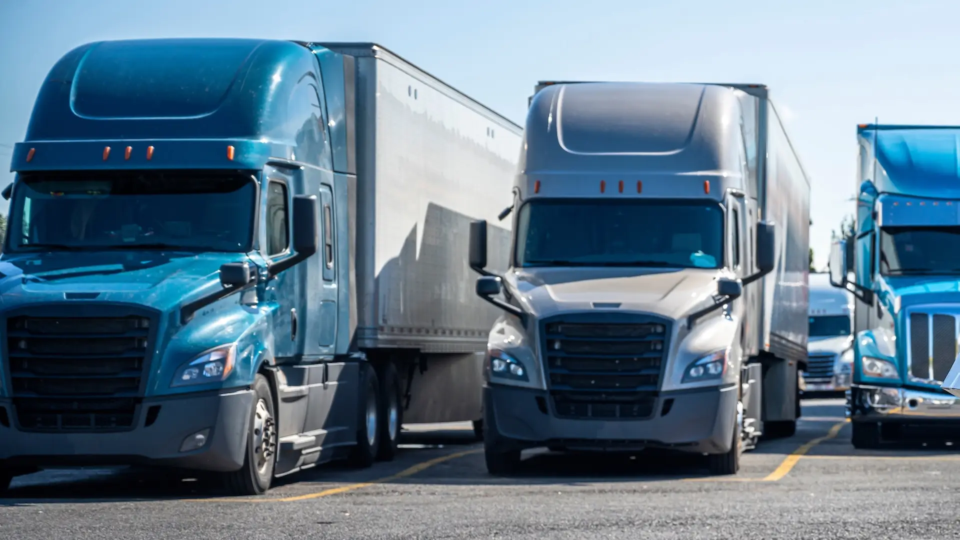 Three semi-trucks in teal, tan, and bright blue park side-by-side in a lot under a clear sky.