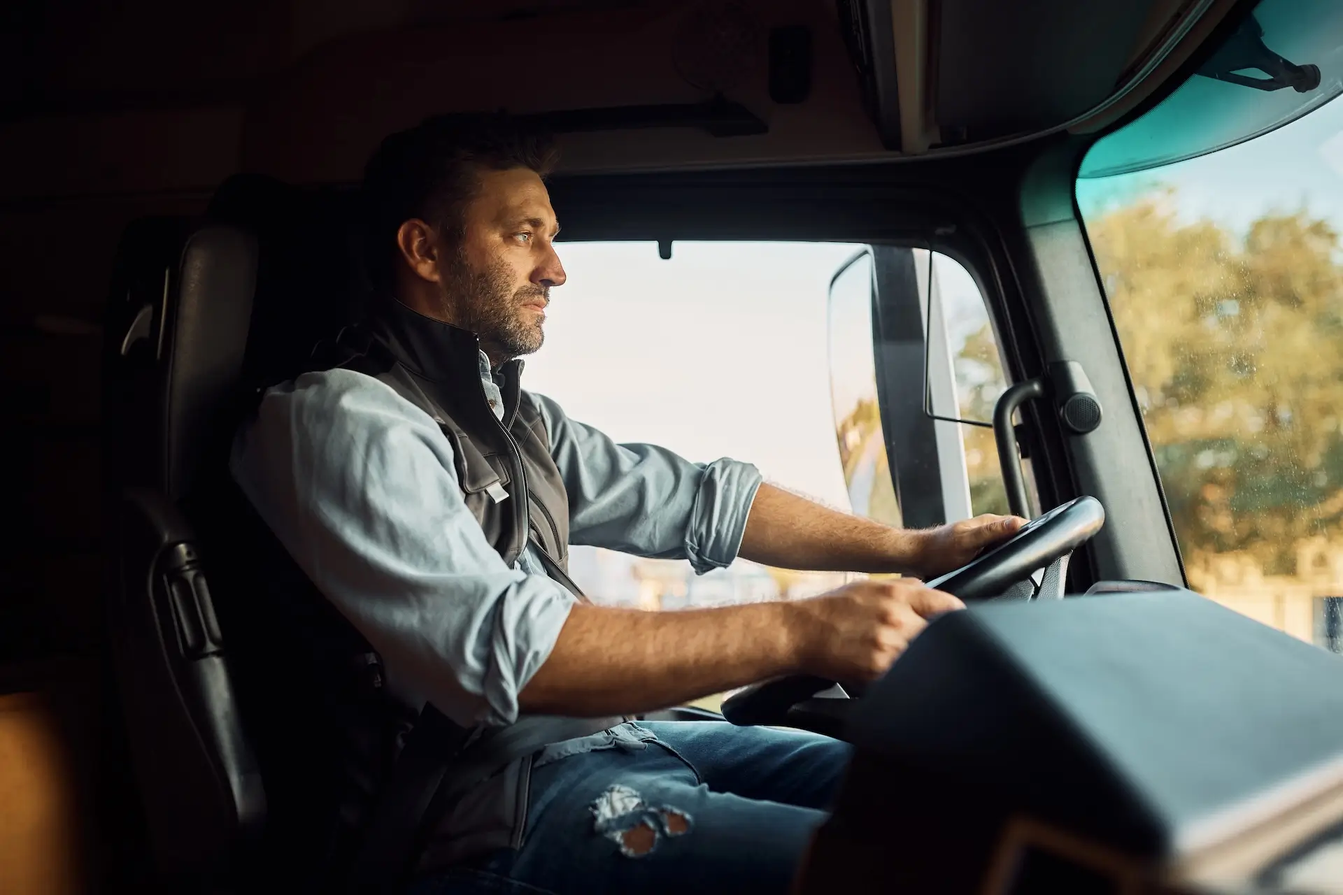 A man with a medium-light skin tone and light facial hair drives a vehicle, looking ahead with a focused expression. He wears a light blue shirt and a dark vest.