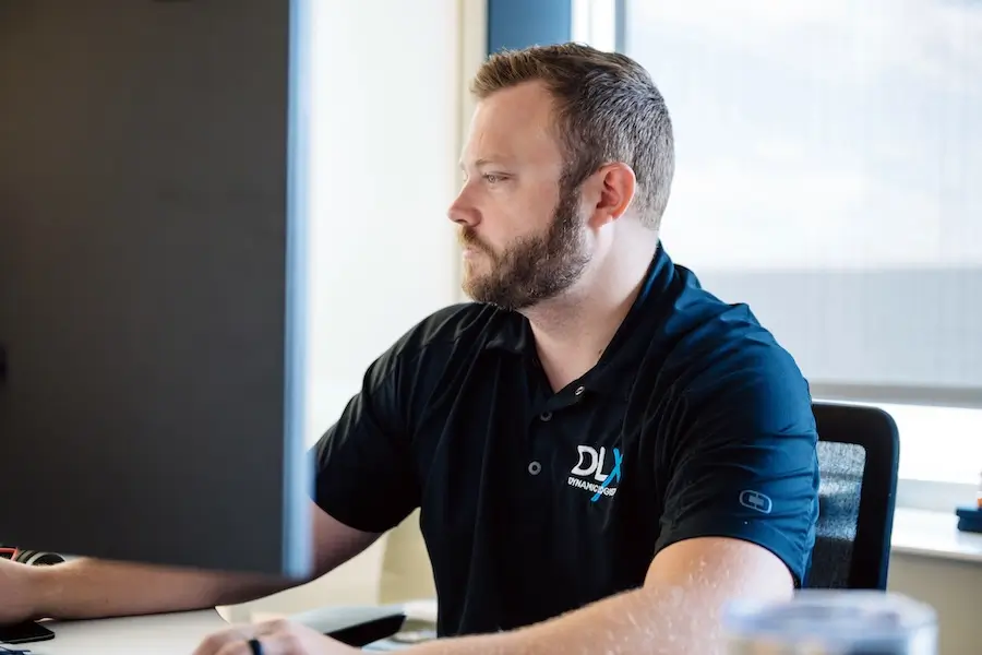 A person with a light skin tone and a beard wearing a black DLX polo shirt sits at a desk and looks at a computer monitor.