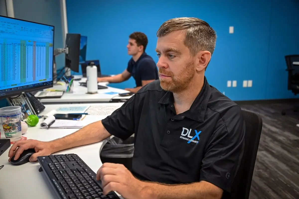 Two people with light skin tones sit at desks in an office with blue walls, focused on computer monitors. The individual in the foreground has short brown hair and a beard, and wears a black polo shirt with the DLX Dynamic Logistics logo.