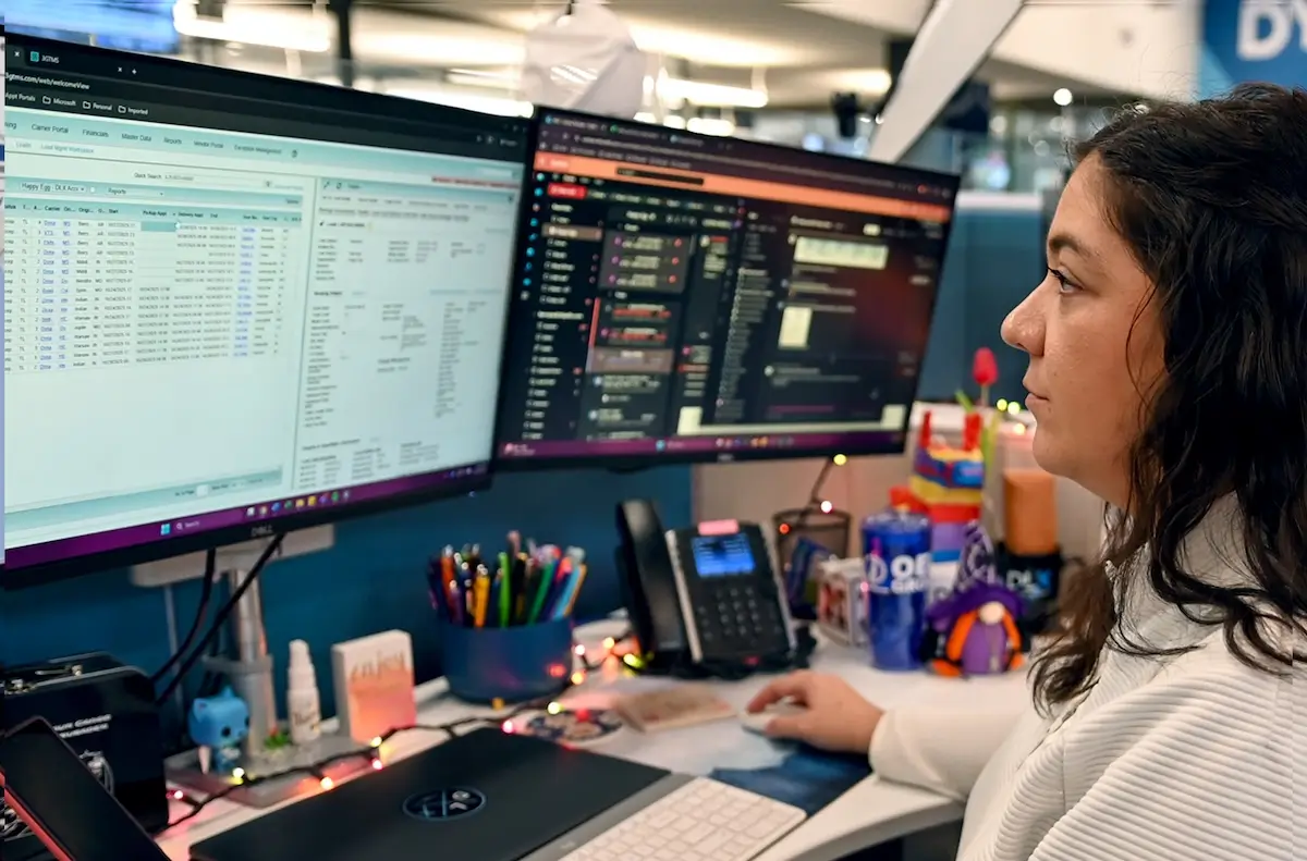 A person with light skin tone and long dark hair looks at two computer monitors displaying data spreadsheets while working at a desk.