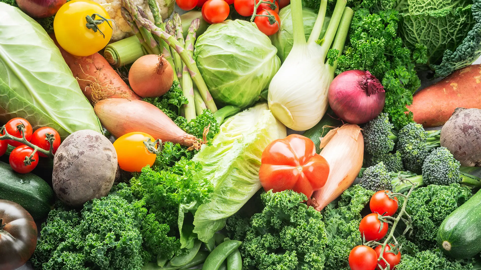 A vibrant, overhead arrangement of various fresh vegetables, including kale, tomatoes, onions, broccoli, and cabbage.